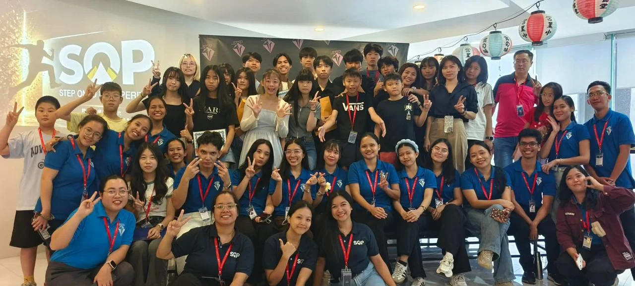 SOP 國際語言學校師生大合照，數十位學員與身穿 SOP 制服的教師在校內 SOP STEP ONTO PROSPERITY 品牌牆前合影